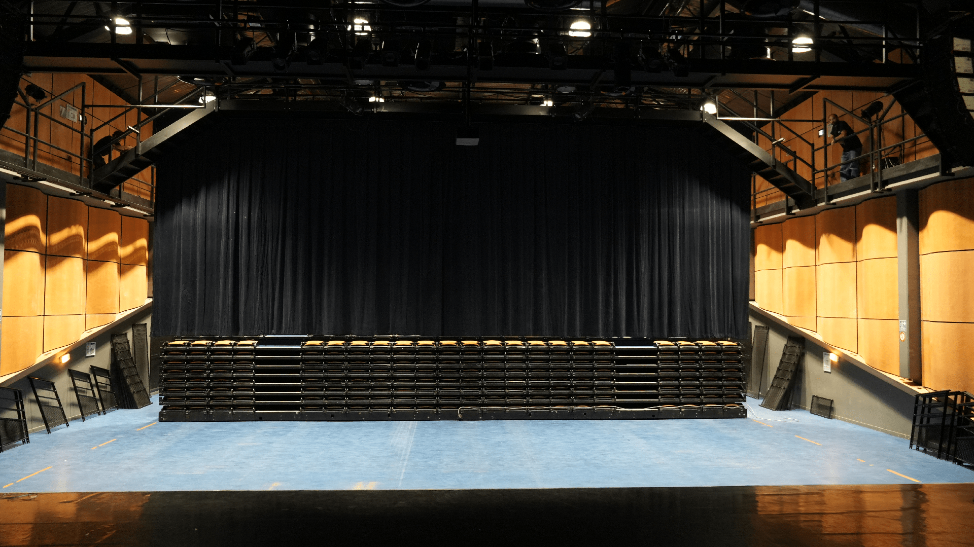 Salle de spectacle vide avec gradins repliés et un grand rideau noir au fond.