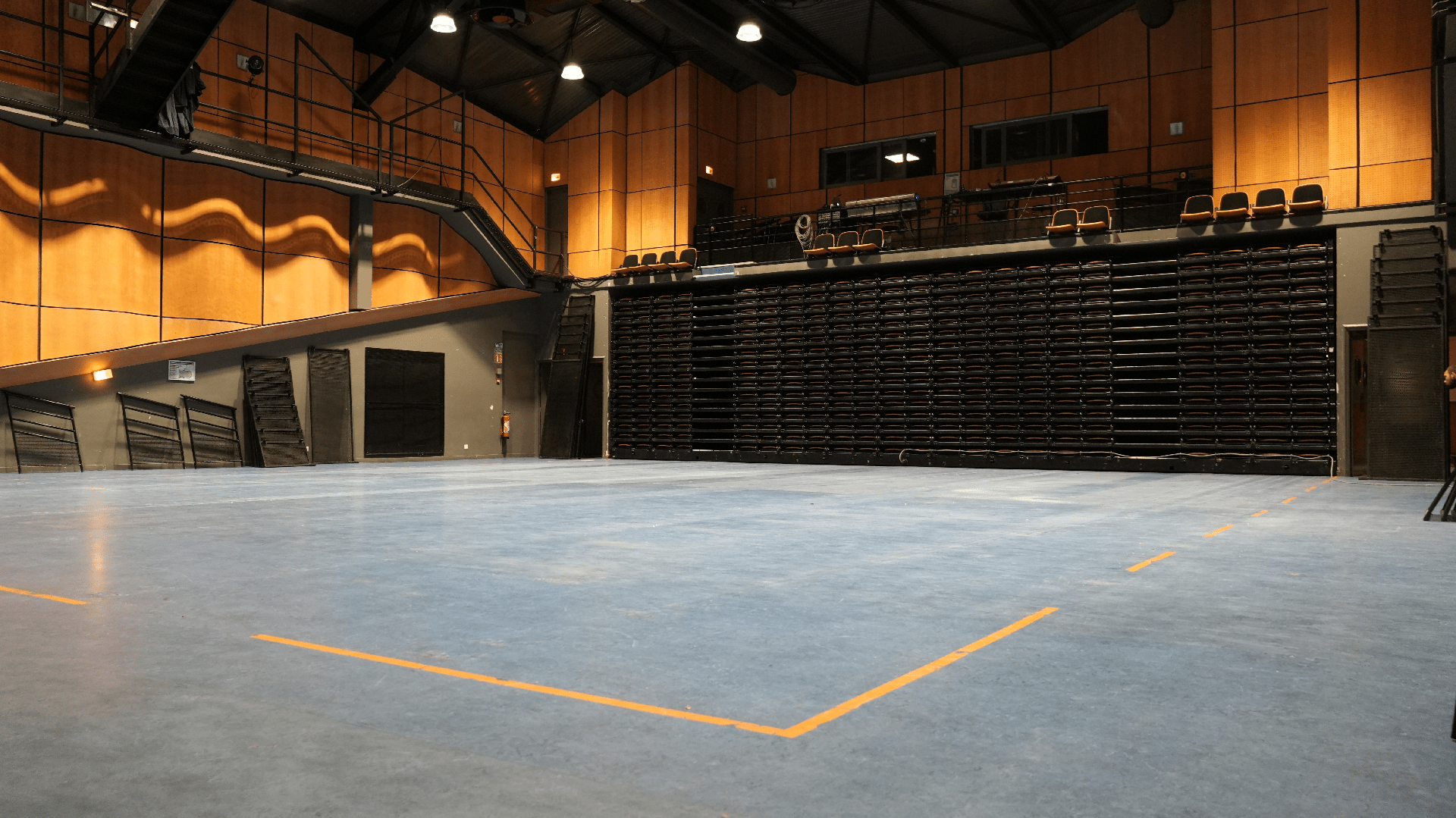 Grande salle vide avec gradins entièrement repliés et sol bleu marqué de lignes orange.