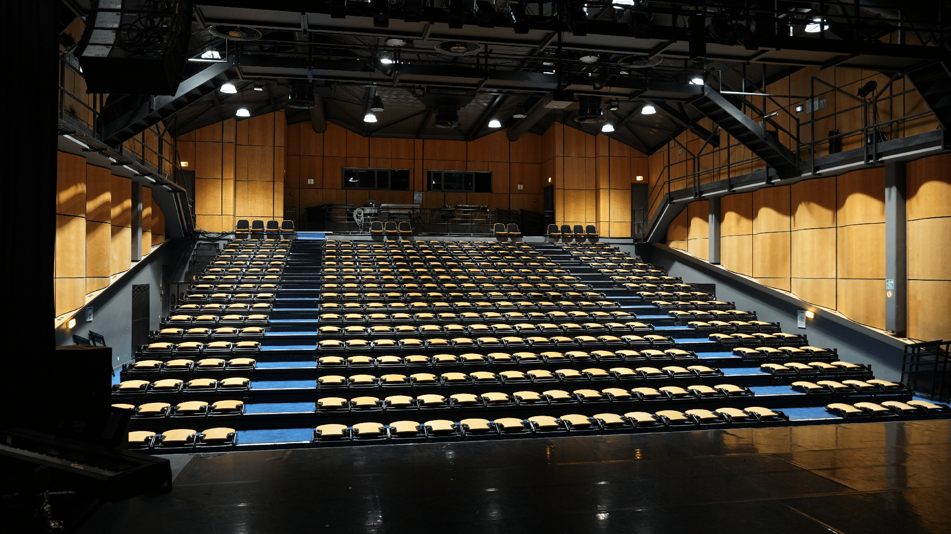 Salle de spectacle vide vue depuis la scène, avec gradins de sièges jaunes et structure métallique au plafond.